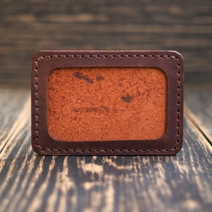 May include: A brown leather card holder with a stitched border and a blank interior.