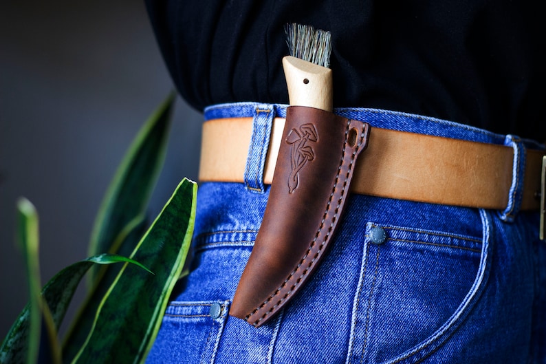 K&ouml;nnte beinhalten: Ein braunes Ledermesser-Etui mit einem Pilz-Design, das an einem braunen Lederg&uuml;rtel &uuml;ber einer blauen Jeans getragen wird.