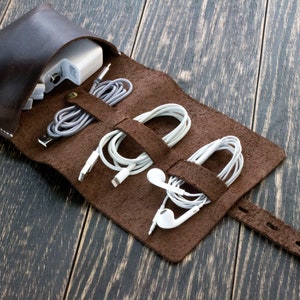 May include: A brown leather roll-up case with three compartments for storing cords and headphones. The case has a strap with a buckle closure.