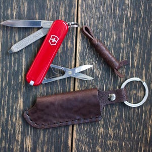 May include: A red Swiss Army knife with multiple tools, including a blade, nail file, and scissors, is displayed open. A brown leather sheath with a keyring and a matching leather lanyard are also shown. The items are arranged on a dark wood surface.