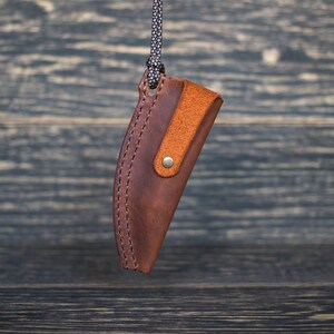 K&ouml;nnte beinhalten: Eine braune Ledertasche f&uuml;r ein Messer mit einem braunen Lederriemen und einem Messingverschluss. Die Tasche h&auml;ngt an einer schwarzen Schnur.