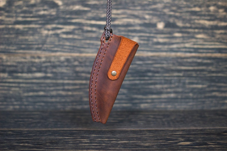 K&ouml;nnte beinhalten: Eine braune Ledertasche f&uuml;r ein Messer mit einem braunen Lederriemen und einem Messingverschluss. Die Tasche h&auml;ngt an einer schwarzen Schnur.