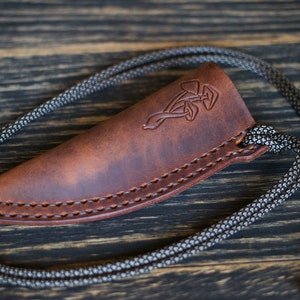 K&ouml;nnte beinhalten: Eine braune Ledertasche f&uuml;r ein Messer mit einem dekorativen keltischen Knotendesign. Die Tasche hat eine schwarze Schnur.