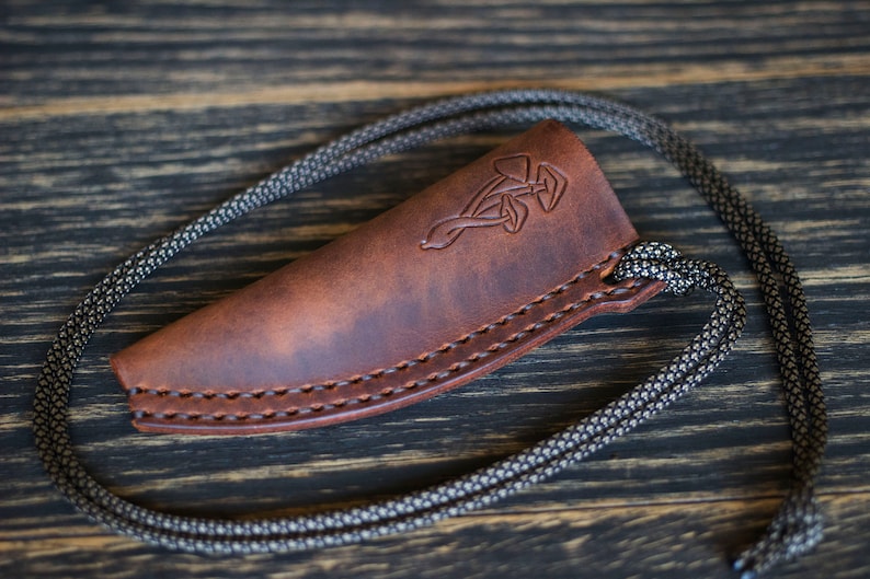 K&ouml;nnte beinhalten: Eine braune Ledertasche f&uuml;r ein Messer mit einem dekorativen keltischen Knotendesign. Die Tasche hat eine schwarze Schnur.