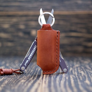 Op de afbeelding: Een bruin leren etui met een zilveren multitool erin. Het etui heeft een zilveren ring aan de bovenkant en de multitool heeft de tekst "Leatherman Micra" erop gedrukt.