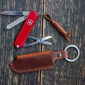 Puede incluir: Una navaja suiza roja con múltiples herramientas, incluyendo una hoja y tijeras, junto a un llavero de cuero marrón y una funda de cuero. Los artículos están dispuestos sobre una superficie de madera oscura.