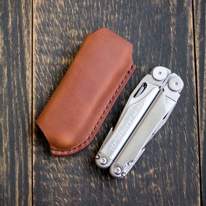 Peut inclure: Un outil multifonction Leatherman Wave argenté en position repliée avec la marque gravée sur le manche, accompagné d'un étui en cuir marron doté de coutures visibles sur les bords et d'un rabat supérieur arrondi.