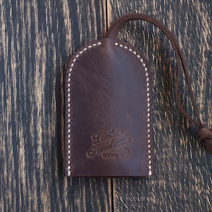 Op de afbeelding: Een bruine leren sleutelhanger met witte stiksels en de tekst "Leather Handmades EST.2013" gegraveerd op de voorkant.