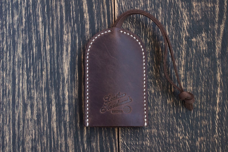 Op de afbeelding: Een bruine leren sleutelhanger met witte stiksels en de tekst "Leather Handmades EST.2013" gegraveerd op de voorkant.