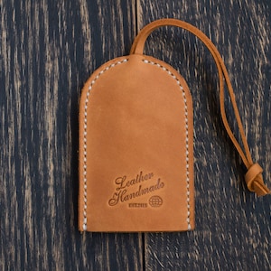 Op de afbeelding: Een bruine leren sleutelhanger met een gestikte rand en een lus. De sleutelhanger heeft de tekst "Leather Handmade" en een wereldbol symbool.