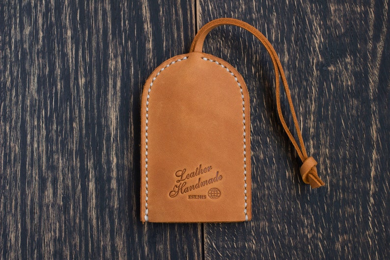 Op de afbeelding: Een bruine leren sleutelhanger met een gestikte rand en een lus. De sleutelhanger heeft de tekst "Leather Handmade" en een wereldbol symbool.
