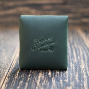 Pode incluir: Uma bolsa de couro verde escura com o texto "Leather Handmade" e um logotipo de globo terrestre em relevo na frente.