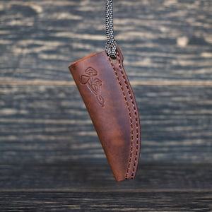 K&ouml;nnte beinhalten: Eine braune Ledertasche f&uuml;r ein Messer mit einem dekorativen Design aus Pilzen und einem keltischen Knoten. Die Tasche hat einen gen&auml;hten Rand und eine Schlaufe f&uuml;r eine Schnur oben.