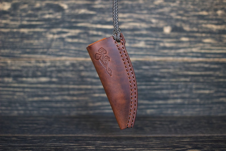 K&ouml;nnte beinhalten: Eine braune Ledertasche f&uuml;r ein Messer mit einem dekorativen Design aus Pilzen und einem keltischen Knoten. Die Tasche hat einen gen&auml;hten Rand und eine Schlaufe f&uuml;r eine Schnur oben.