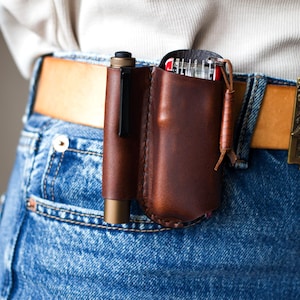 Pode incluir: Um organizador de couro castanho montado no cinto com uma caneta e uma multi-ferramenta. O organizador está preso a um cinto de couro castanho usado com jeans azuis. Uma fivela decorativa com uma caveira é visível.