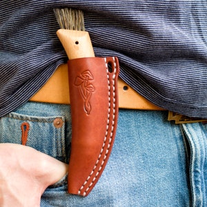 Puede incluir: Una funda de cuchillo de cuero marrón con costuras blancas y un diseño floral. La funda está unida a un cinturón de cuero marrón.