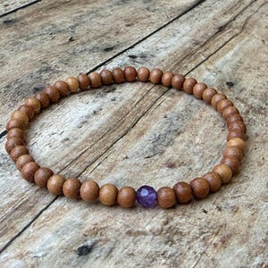 May include: A circular stretch bracelet composed of numerous small, smooth, light brown wooden beads. A single, faceted, translucent purple gemstone bead is positioned as a focal point within the continuous loop of wooden beads.