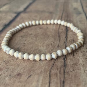 May include: A simple wooden bracelet made of small, round beads. The beads are a light beige color and are strung together on a thin cord.