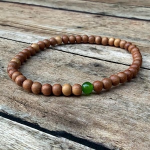 May include: A circular beaded bracelet composed of numerous small, smooth, light brown wooden beads arranged in a continuous loop, featuring a single, polished, vibrant green spherical bead positioned as a distinct accent within the strand.