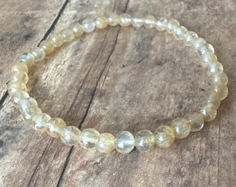 May include: A beaded bracelet composed of small, round, translucent yellow-orange gemstones. The bracelet is strung on an elastic band and rests on a wooden surface. The gemstones have a slightly faceted appearance.