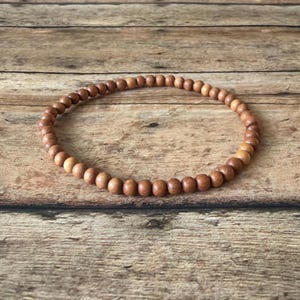 May include: A circular bracelet composed of small, uniform, round wooden beads in a natural light brown tone. The beads are strung together in a continuous loop, showcasing a smooth, matte texture and consistent grain pattern throughout the piece.