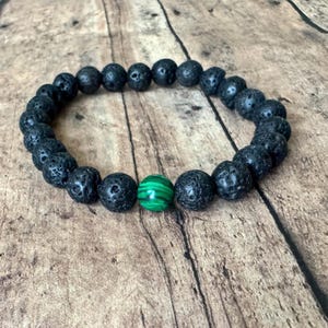 May include: A black lava stone bead bracelet with a single green malachite bead. The bracelet is made of round, textured black beads and a single, polished green bead with a striped pattern. The bracelet is displayed on a wooden surface.