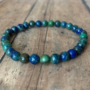 May include: A beaded bracelet featuring a mix of blue and green spherical beads. The beads have a marbled appearance, with varying shades of blue and green. The bracelet is displayed on a wooden surface.