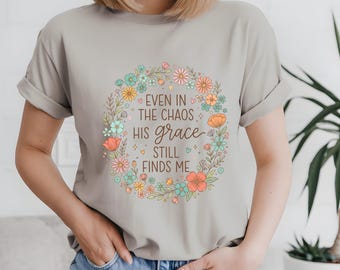 Peut inclure: T-shirt gris clair avec une couronne florale et le texte "EVEN IN THE CHAOS HIS GRACE STILL FINDS ME". La couronne comprend des fleurs roses, bleues et oranges.