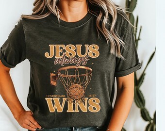 Peut inclure: T-shirt vert foncé avec un motif de panier de basket et le texte doré "JESUS always WINS". Le design comprend un ballon de basket et le verset biblique 1 Corinthiens 15:57.