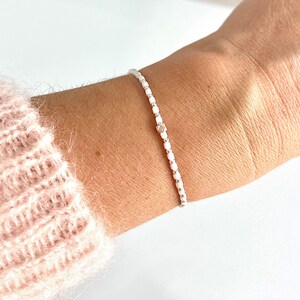 Peut inclure: Un bracelet en perles blanches avec un charme argenté. Le bracelet est porté au poignet.