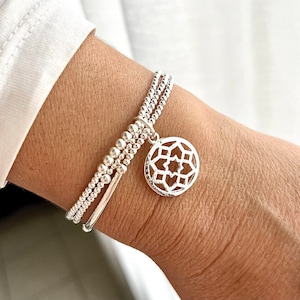 Könnte beinhalten: Ein silbernes Armband mit einer zarten Kette und einem runden Anhänger mit geometrischem Muster. Das Armband ist mit kleinen silbernen Perlen geschichtet.