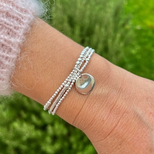 Op de afbeelding: Een zilveren armband met drie rijen kleine kralen en een ronde hanger. De armband wordt om de pols gedragen.