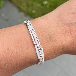 Peut inclure: Un ensemble de trois bracelets en argent avec des perles blanches. Les bracelets sont empilés sur le poignet d'une personne.