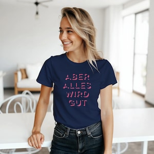 May include: Navy blue t-shirt with the pink text "ABER ALLES WIRD GUT". The shirt is a crew neck style and is made of a soft material. The shirt is worn by a person.