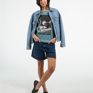 Peut inclure: Une femme portant un t-shirt bleu clair avec un imprimé graphique noir et blanc d'une personne courant avec le texte "INTERCEPTING NAPOLEON". Elle porte également une veste en jean et un short en jean bleu foncé.