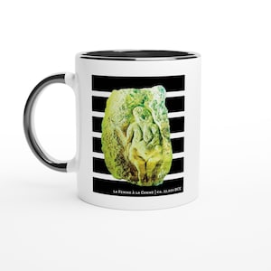 Puede incluir: Taza de cerámica blanca con interior y asa negros. La taza presenta una imagen verde y amarilla de una escultura sobre un fondo de rayas blancas y negras. El texto "LA FEMME À LA CORNE | CA. 23.000 a. C." está impreso en la parte inferior.