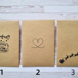 May include: Three brown paper envelopes with different designs. The first envelope has a black and white illustration of a house with a Christmas tree and the text "Happy Holidays". The second envelope has a black and white illustration of a heart. The third envelope has a black and white illustration of a reindeer pulling a sleigh.