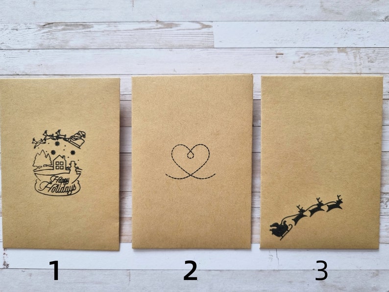 May include: Three brown paper envelopes with different designs. The first envelope has a black and white illustration of a house with a Christmas tree and the text "Happy Holidays". The second envelope has a black and white illustration of a heart. The third envelope has a black and white illustration of a reindeer pulling a sleigh.