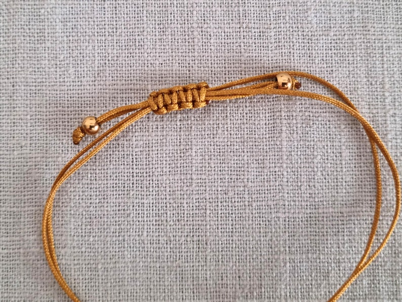 May include: A gold cord bracelet with two small gold beads. The cord is knotted in a decorative pattern.