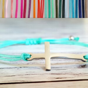 Könnte beinhalten: Ein türkisfarbenes Armband aus Kordel mit einem silbernen Kreuzanhänger. Das Armband ist verstellbar und kann sowohl von Männern als auch von Frauen getragen werden.