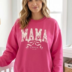 May include: A bright pink sweatshirt with the word "MAMA" in a floral design. Below the word "MAMA" is a bow and the name "Laura" with the text "est. 2021". The sweatshirt is a crew neck style.