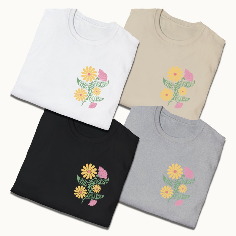 May include: Four folded t-shirts in white, beige, black, and gray with a floral design and the text "The Trust Process" printed on the front.