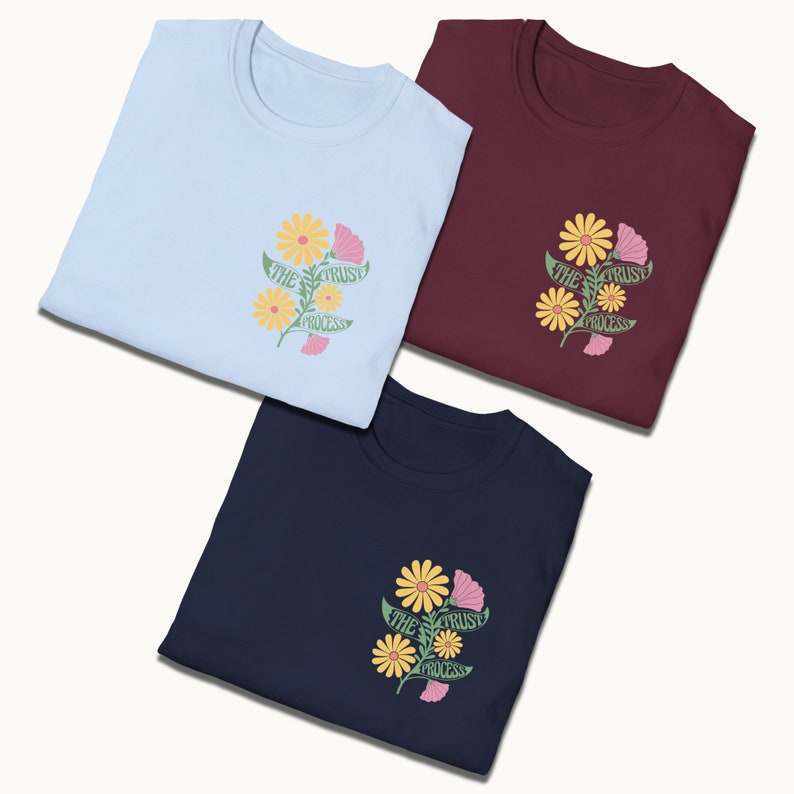 May include: Three folded t-shirts in different colors: light blue, maroon, and navy blue. Each shirt features a floral design with yellow and pink flowers and green leaves. The text "The Trust Process" is written on the flowers.