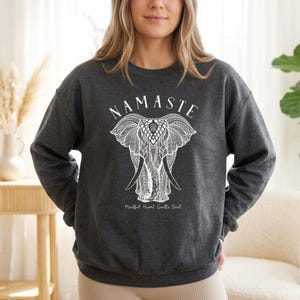 Puede incluir: Sudadera gris oscuro con un diseño de elefante blanco y la palabra "NAMASTE" encima. El elefante está intrincadamente detallado con un patrón de estilo mandala. Debajo del elefante está el texto "Mindful Heart Gentle Soul".