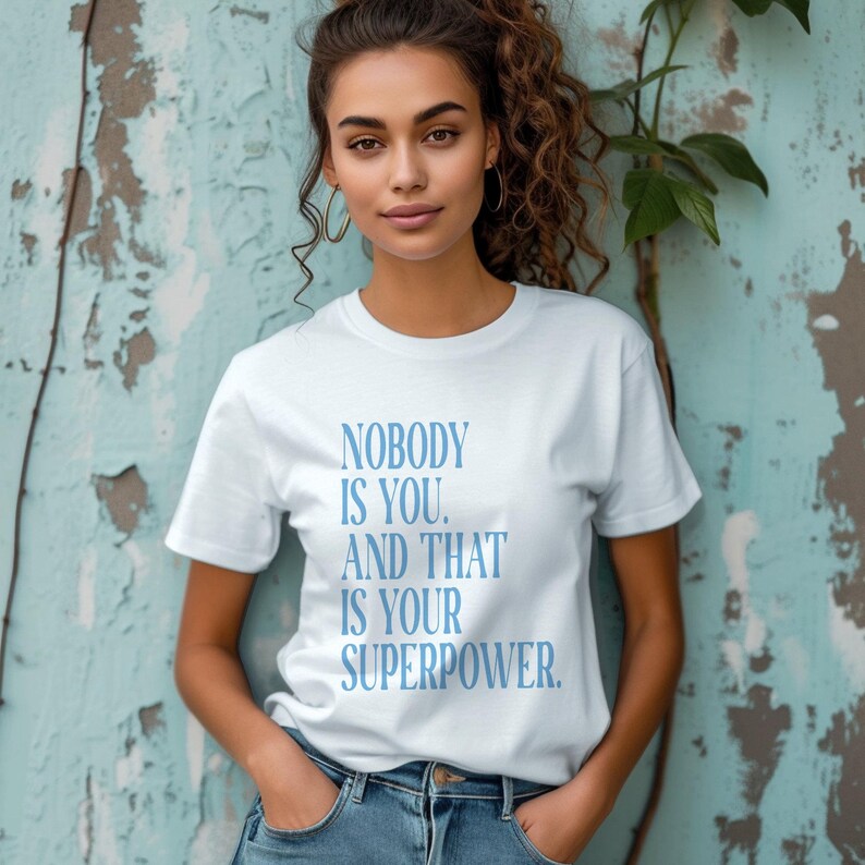 May include: White t-shirt with the text "NOBODY IS YOU. AND THAT IS YOUR SUPERPOWER." in light blue. The shirt is a crew neck style and is being worn.