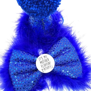 May include: A blue sequined bow with a white button that says "It's my mother fluffin' birthday!" The bow is surrounded by blue feathers.