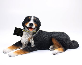 May include: A black, white, and brown Bernese Mountain Dog figurine with a yellow collar and a silver tag that says "Buddy". The figurine is lying down with its head turned to the side and its mouth slightly open.