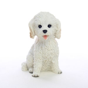 May include: White ceramic figurine of a small dog with fluffy white fur, black eyes, and a pink tongue sticking out. The dog is sitting with its legs crossed and its head tilted slightly to the side.