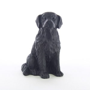 May include: Black Labrador Retriever figurine, sitting with its paws tucked underneath, in a realistic pose. The figurine is made of a dark, matte material.