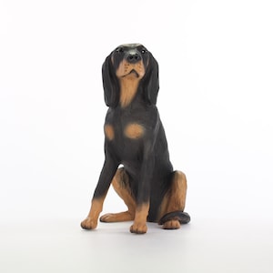 May include: A black and tan figurine of a dog sitting with its head tilted up. The dog has a short, smooth coat and a long tail.